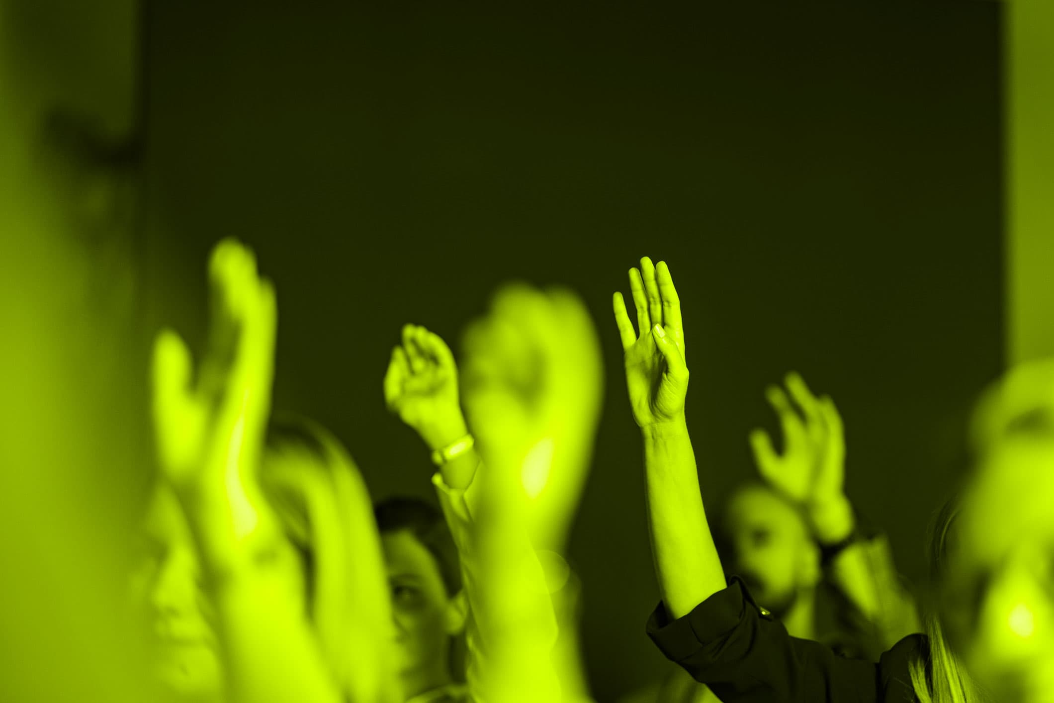 A group of people raise their hands in a dimly lit room. The image is tinted with a green hue.