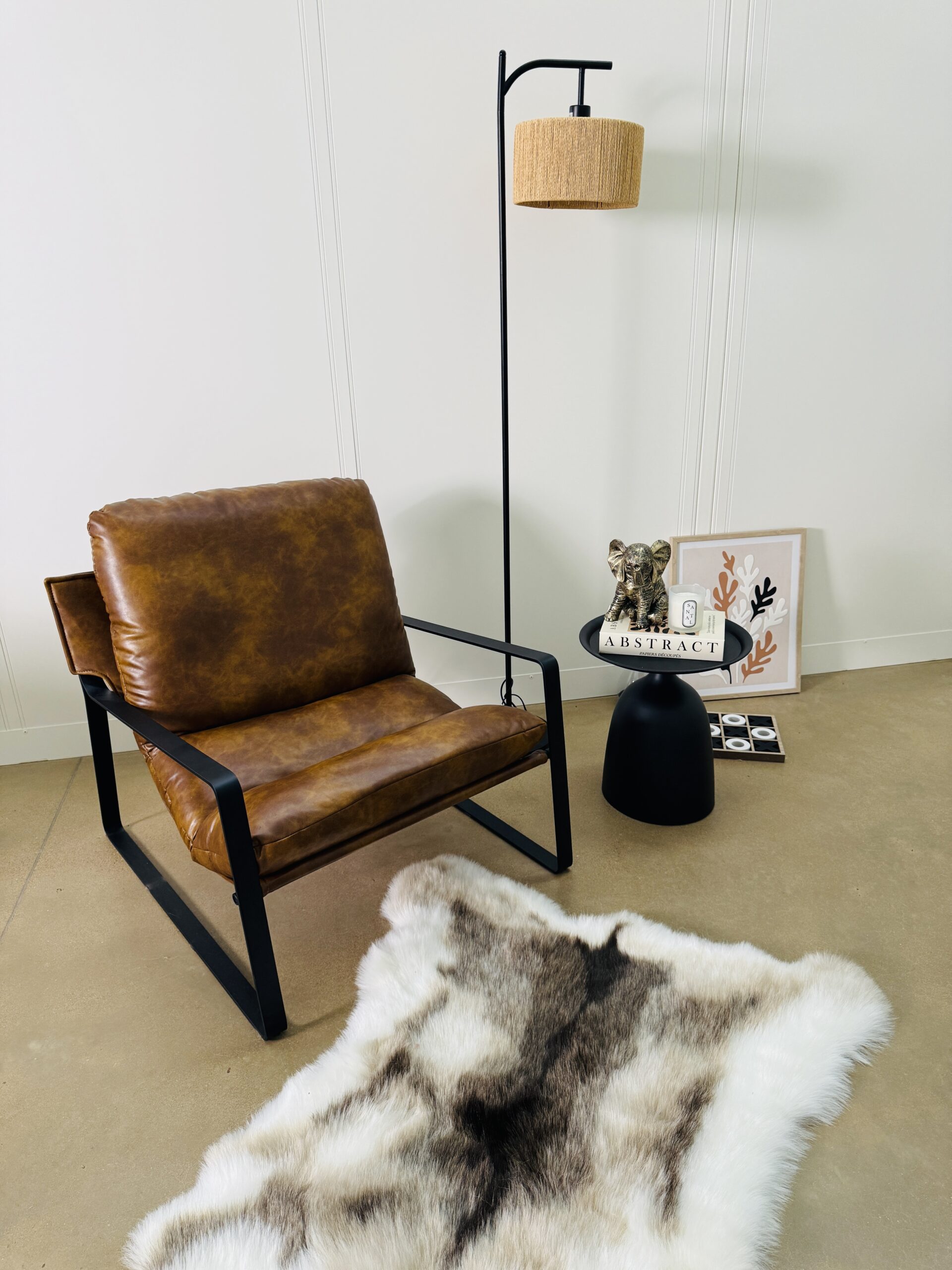 A cozy living space featuring a brown leather armchair beside a black side table topped with a decorative object, a book titled “Abstract,” and a candle. A black floor lamp with a woven wicker shade stands nearby, next to a soft faux-fur rug on beige carpet, with a decorative art print displayed behind the table.