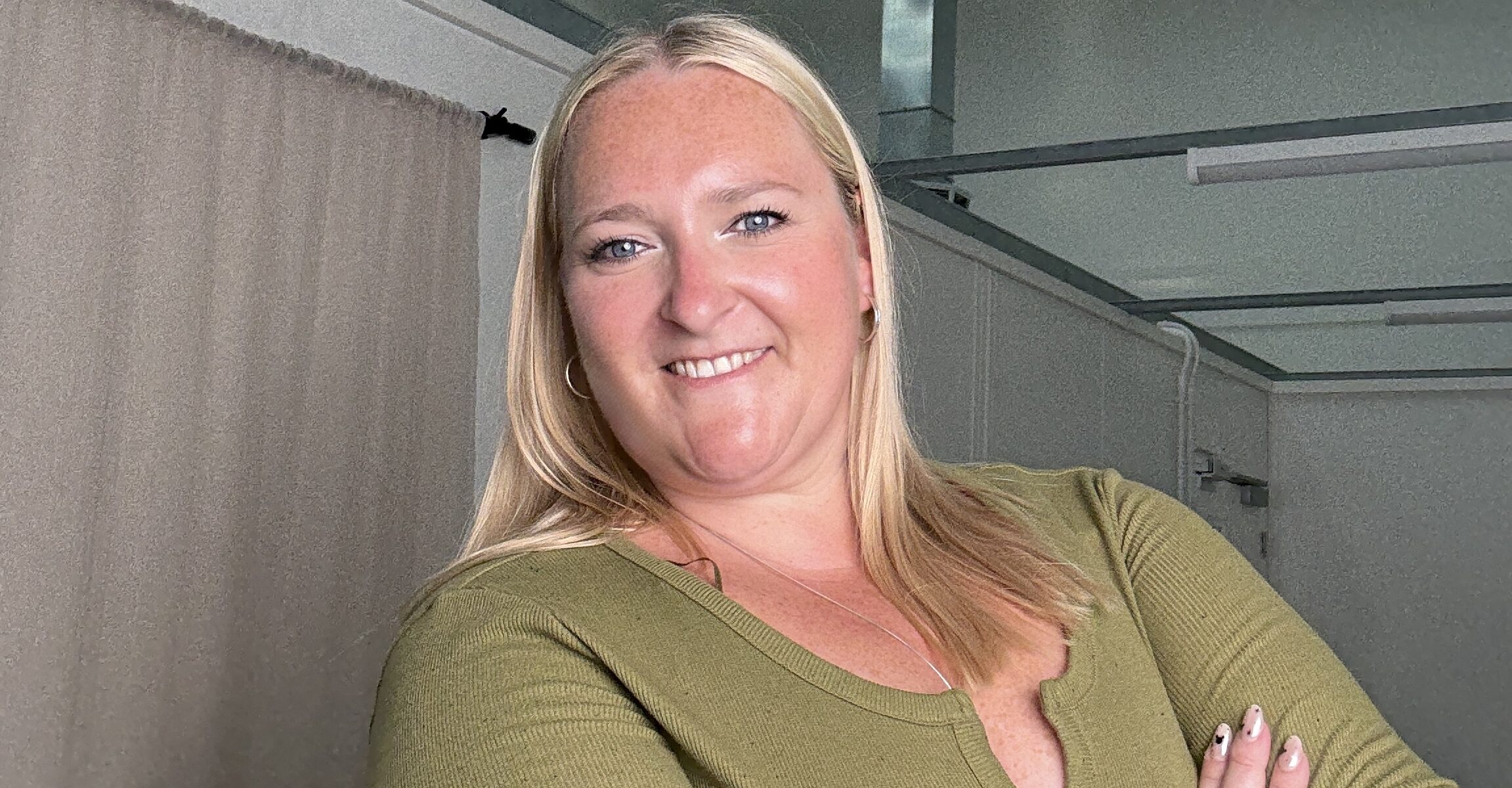 A zoomed-in picture of Kerry Pitt, dressed in a green top, stands with her arms crossed and smiles confidently toward the camera in a softly lit, neutral-coloured space.