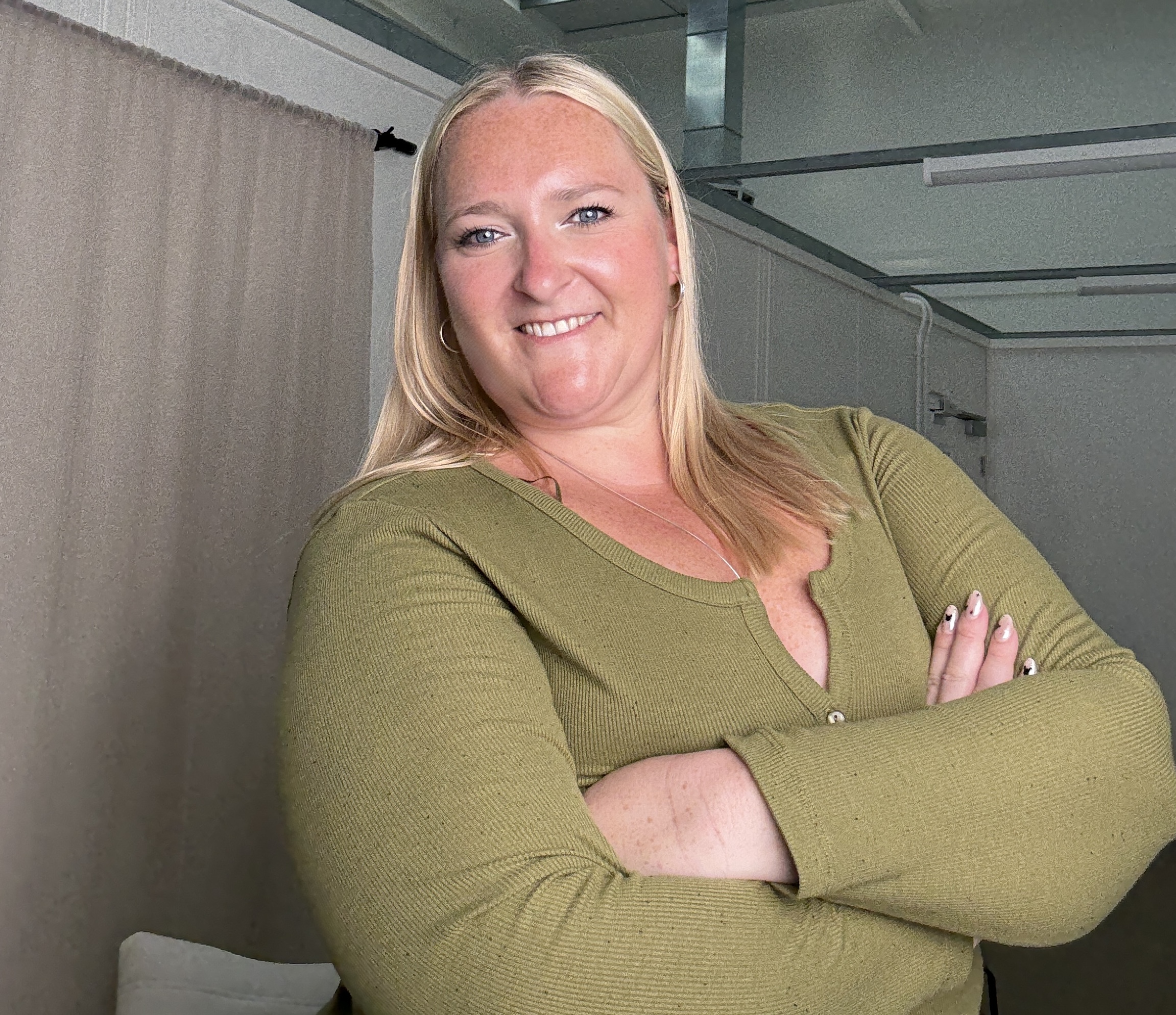 A picture of Kerry Pitt, dressed in a green top, stands with her arms crossed and smiles confidently toward the camera in a softly lit, neutral-coloured space with beige curtains.