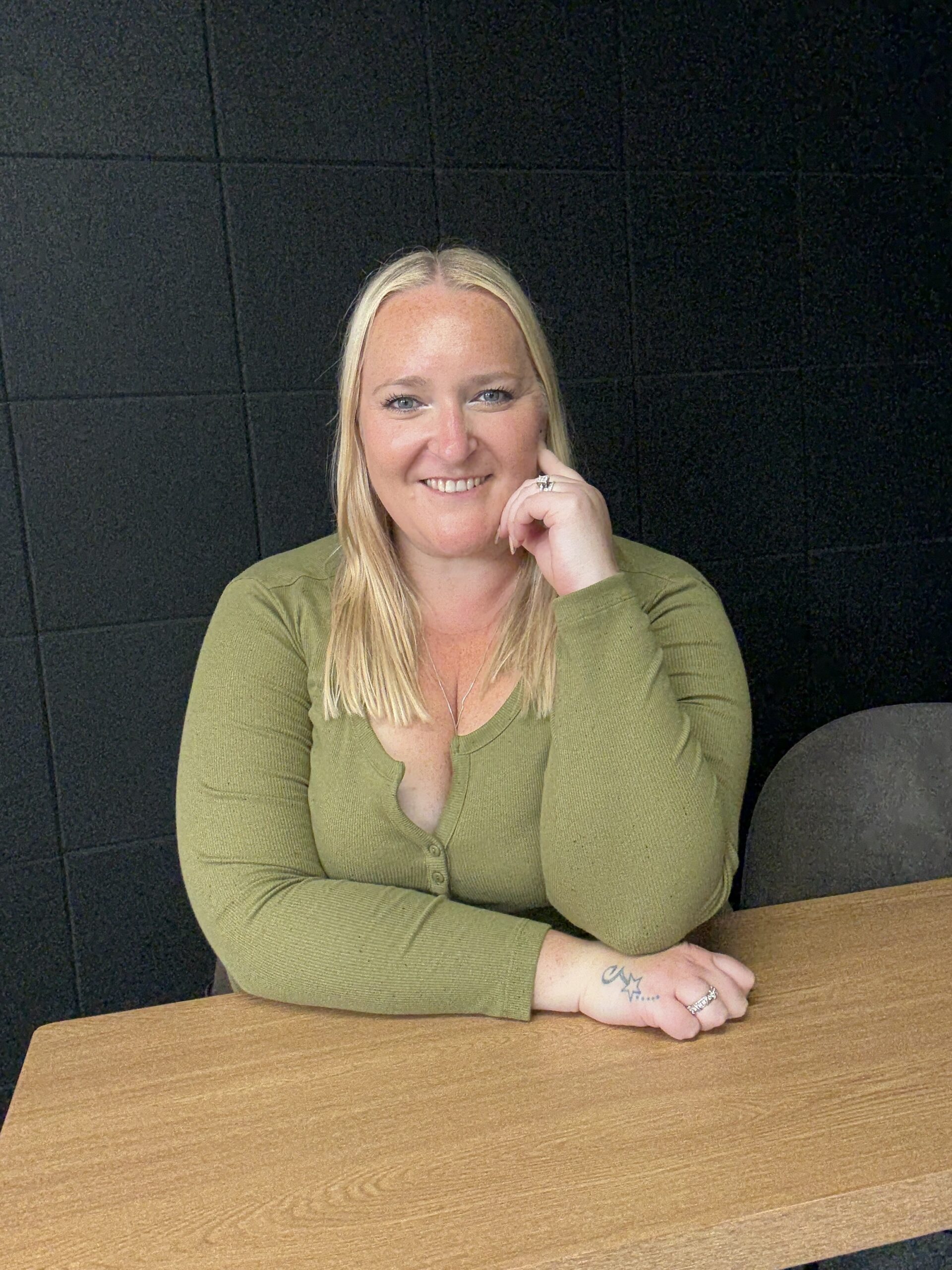 Kerry Pitt sits at a wooden desk, leaning her head on her hand and smiling as she to the camera. She is wearing a green top, with a dark, tiled square-patterned background behind her.