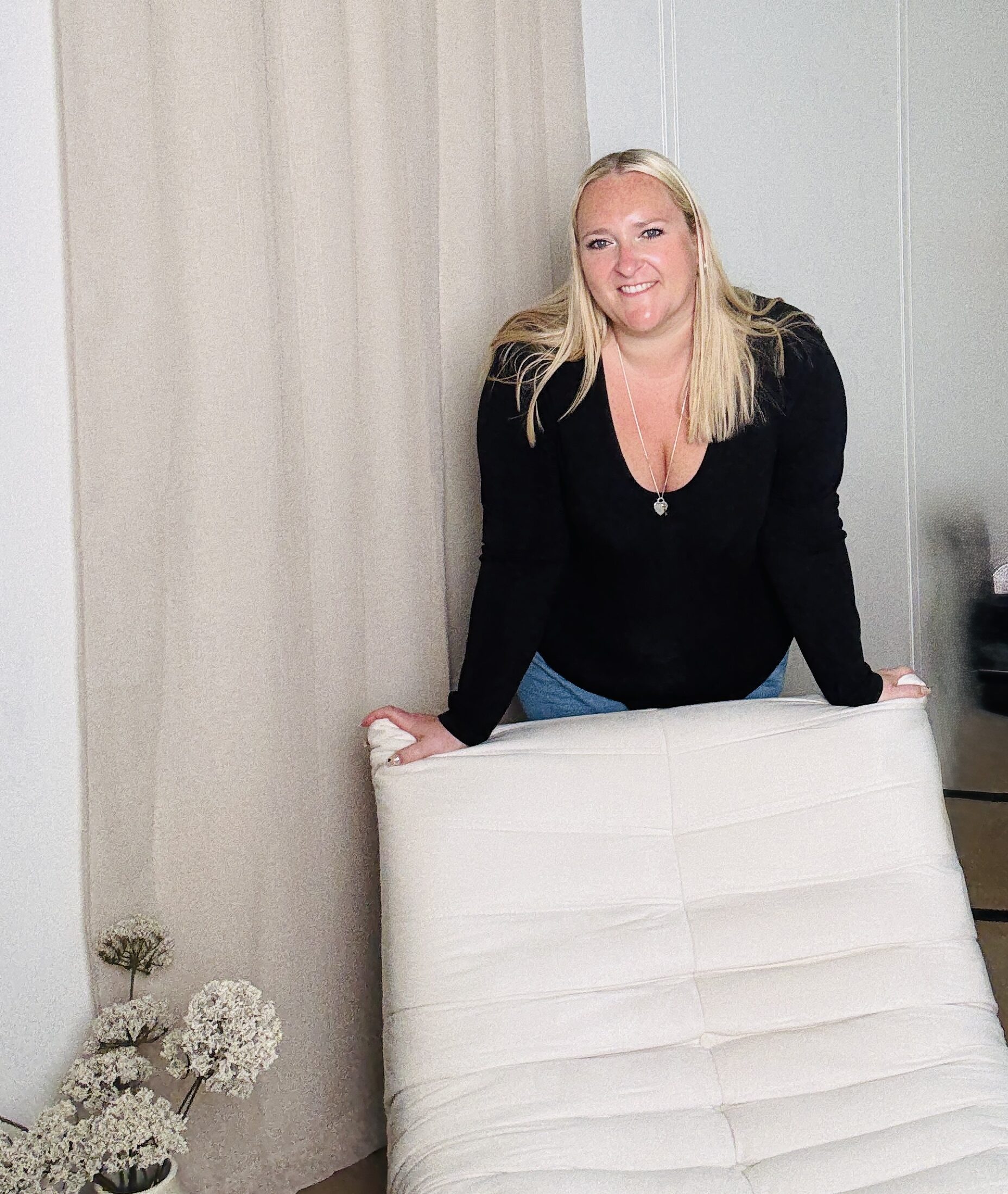 A slightly zoomed picture of Kerry Pitt smiling warmly in a cosy, neutral-toned room framed by soft beige curtains, leaning over the back of a cream-coloured chair. She is dressed in a black top and blue jeans.