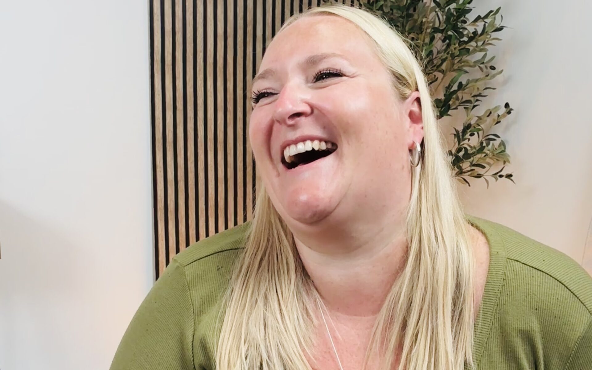 A close-up image of Kerry Pitt, wearing a green top, sits laughing while facing to the left, with vertical wall panelling and a leafy plant in the background.